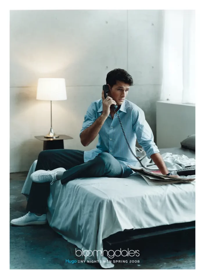 A male model wearing a Hugo Boss striped shirt, sitting on a bed with a telephone. Campaign by PlanetFab Studio
