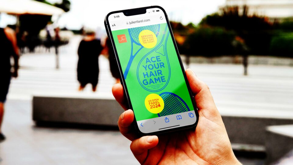 Smartphone displaying Julien Farel's brand campaign US Open 2024 'Ace Your Hair Game' promotion website, held against blurred urban background
