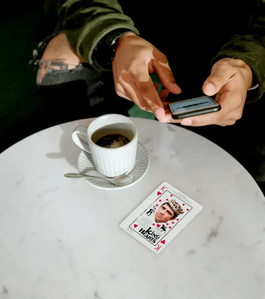 Hands using smartphone above coffee cup and King of Hearts key art poster design on marble table. King of Hearts movie newspaper ad. Key Art poster design and campaign by PlanetFab Studio.