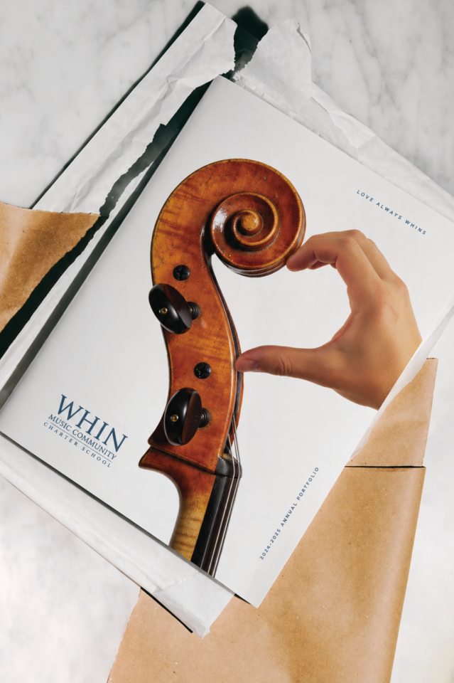 WHIN Music Community Charter School 2024-2025 Annual Portfolio cover, featuring a cello scroll with a hand forming a heart shape, styled on marble with kraft and tissue paper.