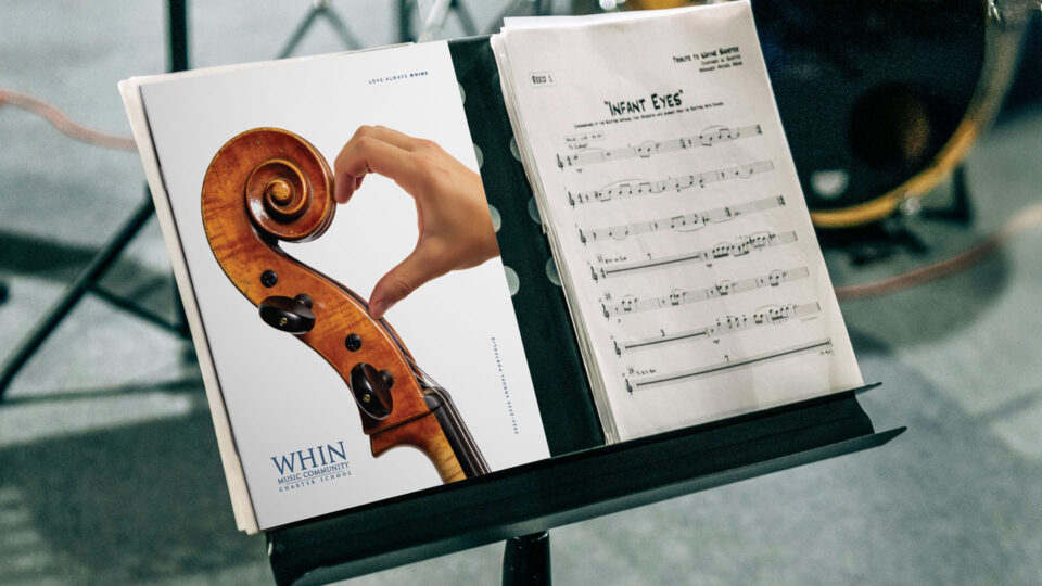 WHIN Music Community Charter School 2024-2025 Annual Portfolio cover on a music stand, featuring a cello scroll paired with a hand forming a heart shape, beside hand-notated sheet music for "Infant Eyes."