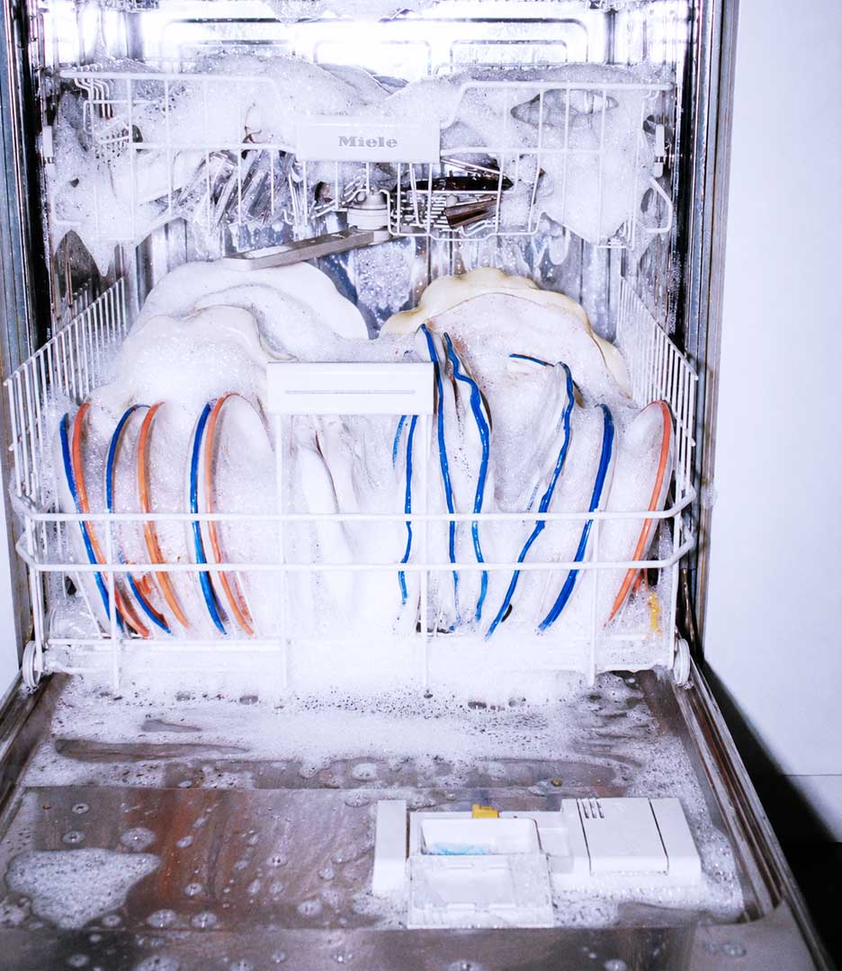 Open dishwasher overflowing with soap suds covering dishes and racks, illustrating a common appliance malfunction. Bernard AI appliance repair platform by PlanetFab.