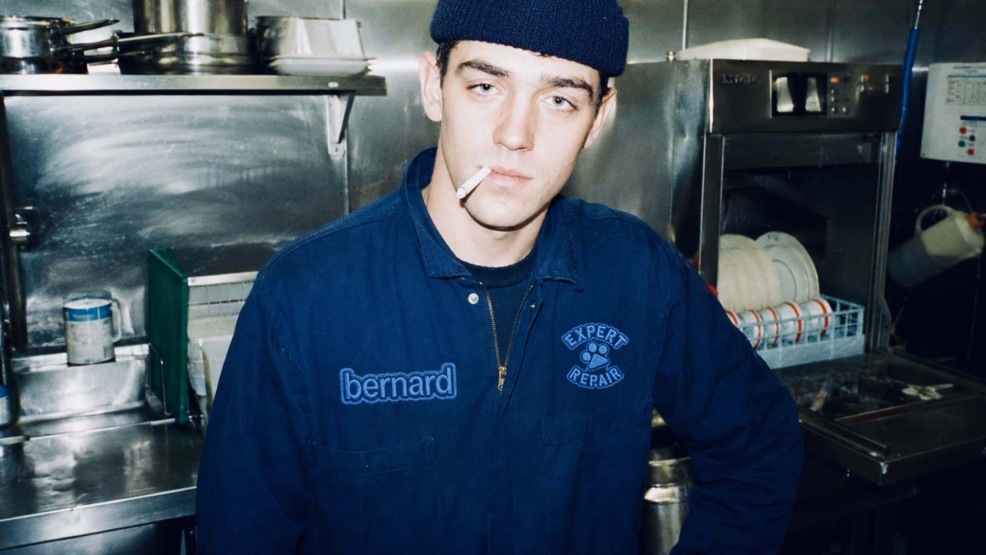 Male technician in a navy work jacket with Bernard name patch and Expert Repair embroidered badge standing in a commercial kitchen. Bernard AI brand identity by PlanetFab.